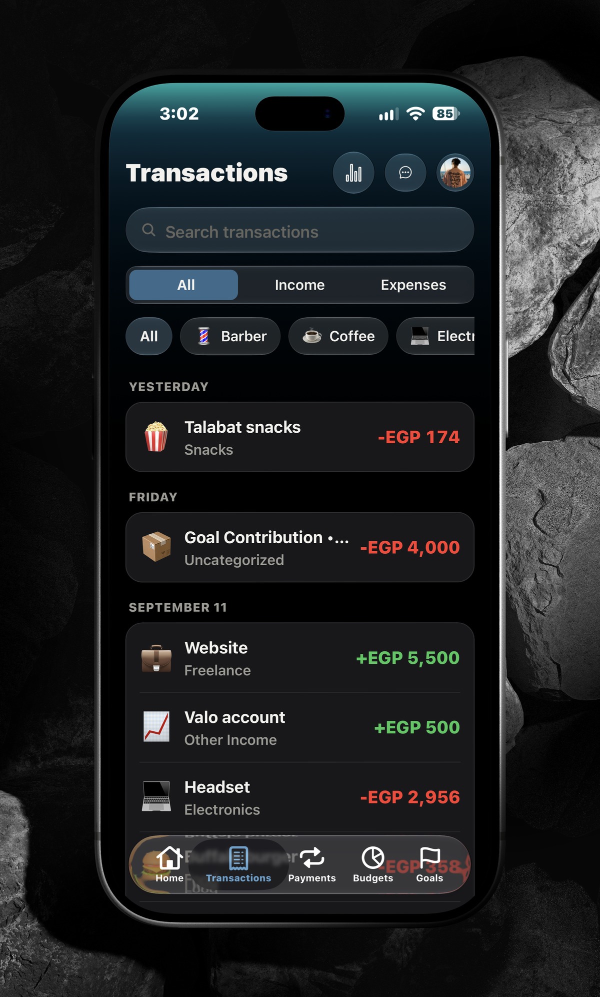 AIVA transactions screen showing a searchable, filterable list of income and expenses grouped by day, on an iPhone lying flat on dark stones